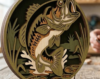 Puede incluir: Una pieza de arte mural circular de madera multicapa que presenta un recorte detallado de un pez lubina saltando. El diseño incorpora tonos verdes, colores de madera natural y marrón, representando al pez entre plantas acuáticas.