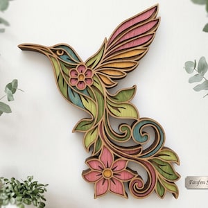 May include: A multi-layered wooden wall art piece shaped like a hummingbird in flight, featuring intricate cutouts, floral motifs, and a color palette of pink, green, yellow, and teal. A small rectangular metal plaque reads Fanfen Studio.