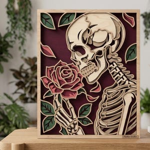 Könnte beinhalten: Ein mehrschichtiges Wandkunstwerk aus Holz mit einem hellen Totenkopf und einer Skeletthand, die eine rosa Rose hält. Das Design umfasst grüne Blätter und verstreute rosa Blütenblätter vor einem tiefroten Hintergrund, gerahmt in hellem Holz.