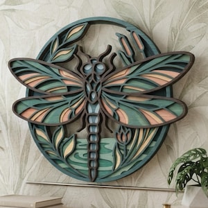 May include: A circular layered wooden wall art piece featuring a central dragonfly design. The artwork uses shades of teal, green, and soft peach, with intricate cutouts depicting wings, cattails, and foliage within a circular frame structure.