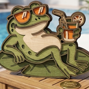 Pode incluir: Uma peça de arte de parede em madeira multicamadas representando um sapo verde com óculos de sol laranja, deitado numa vitória-régia e segurando uma bebida laranja com palhinha e uma fatia de citrino. O texto Fanfen Studio está gravado num disco.