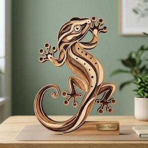 May include: A layered wooden gecko sculpture featuring various shades of brown wood grain. The lizard is depicted in a climbing pose with a curled tail and circular cutouts along its back. It is mounted on a rectangular wooden base with the text Fanfen Studio.