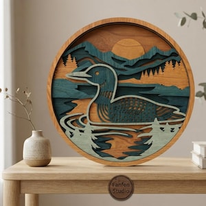 May include: A circular multi-layered wooden wall art piece featuring a loon swimming on water with a mountain and forest background. The design uses shades of blue, green, and natural wood tones. A small circular logo reads Fanfen Studio.