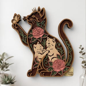May include: A multi-layered wooden wall art piece shaped like a cat reaching for a butterfly. The design features a dark brown cat silhouette containing two light-coloured kittens, pink roses, and green vines. A small gold tag reads Fanfen Studio.