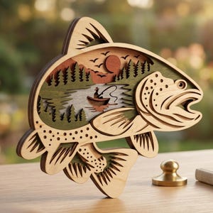 Könnte beinhalten: Ein mehrschichtiger Holzausschnitt in Fischform mit einer lasergeschnittenen Szene einer Person in einem kleinen Boot, die auf einem See angelt. Das Design umfasst Silhouetten von Kiefern, eine kreisförmige Sonne und Vögel.