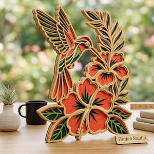 May include: A multi-layered wooden wall art piece featuring a hummingbird hovering over hibiscus flowers. The design uses shades of orange, green, and natural wood tones. A small rectangular plaque at the base displays the text Fanfen Studio.