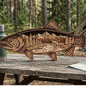 May include: Wooden fish-shaped art piece with a detailed interior scene of mountains, trees, and a jumping fish. The art piece is made of light and dark brown wood and sits on a wooden picnic table. A green thermos is visible.
