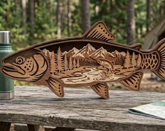 May include: Wooden fish-shaped art piece with a detailed interior scene of mountains, trees, and a jumping fish. The art piece is made of light and dark brown wood and sits on a wooden picnic table. A green thermos is visible.