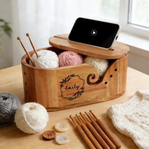 Peut inclure: Une boîte de rangement en bois pour fil à tricoter avec un couvercle, contenant des pelotes de laine blanches et roses. La boîte présente un motif floral avec le nom "Emily". Des aiguilles à tricoter, des crochets et des boutons sont disposés à côté. Un smartphone est posé sur le couvercle.