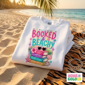 Op de afbeelding: Witte sweatshirt met de tekst "Booked & Beachy" in roze en turquoise. De afbeelding bevat boeken, zonnebrillen, bloemen en een zeester. De sweatshirt is opgevouwen op een strandlaken met tijgerstrepen, met een strand en oceaan op de achtergrond.