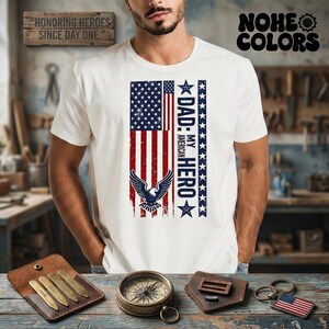 Puede incluir: Camiseta blanca con un diseño de bandera estadounidense desgastada y un águila. El texto "DAD: MY AMERICAN HERO" está impreso verticalmente. La camiseta también tiene el texto "HONORING HEROES SINCE DAY ONE" y "NOHE COLORS".