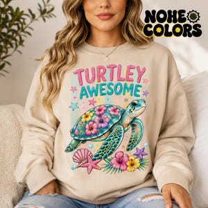 Puede incluir: Sudadera beige con un gráfico colorido de una tortuga marina con caparazón floral, rodeada de estrellas de mar, conchas y flores. El texto "TURTLEY AWESOME" está encima de la tortuga. La marca "NOHE COLORS" está en la esquina superior derecha.
