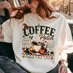 Puede incluir: Una camiseta color hueso con un estampado gráfico que incluye el texto COFFEE Patch. El diseño muestra una taza de café humeante, una pequeña calabaza, un sombrero de bruja, granos de café y hojas de otoño, con PUMPKIN SPICE LATTE en la base.