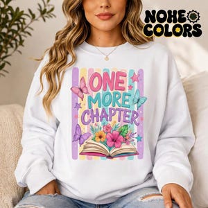 Puede incluir: Sudadera blanca con el texto "ONE MORE CHAPTER" en rosa y azul, decorada con mariposas, estrellas y flores. La sudadera tiene un fondo a rayas de colores y la marca "NOHE COLORS" en la esquina superior derecha.