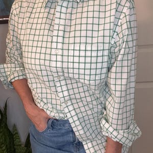 May include: A long-sleeve button-down shirt featuring a white base with a green grid pattern. The shirt includes a standard collar, a single chest pocket on the left side, and white buttons running down the front placket. The sleeves are rolled up.