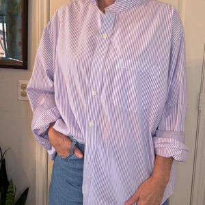 May include: A long-sleeved button-down shirt featuring a vertical lavender and white striped pattern. The shirt includes a ruffled collar, a single chest pocket, and light-colored buttons down the front. The sleeves are rolled up at the cuffs.