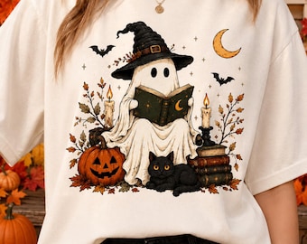 May include: An off-white t-shirt featuring a graphic of a ghost wearing a witch hat and reading a book. The design includes a jack-o'-lantern, a black cat, stacks of books, lit candles, bats, and a crescent moon surrounded by autumn leaves.