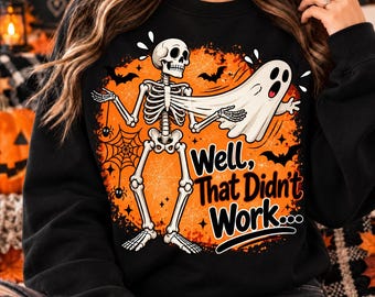 May include: A black sweatshirt featuring a graphic of a white skeleton and a white ghost on an orange circular background. The design includes black bats, spiderwebs, and spiders, with the text "Well, That Didn't Work..." in black lettering.