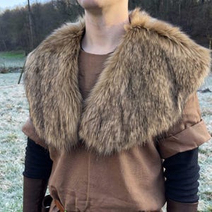 May include: A brown tunic featuring a large, thick faux fur collar in shades of tan and dark brown. The garment includes black long sleeves, brown forearm cuffs, and a brown leather belt around the waist, creating a layered appearance.