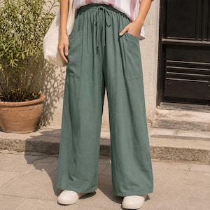 May include: Sage green wide-leg trousers featuring an elasticized waistband with a functional drawstring tie, two large patch pockets on the front, and a loose, flowing silhouette that extends down to the ankles, paired with white sneakers.
