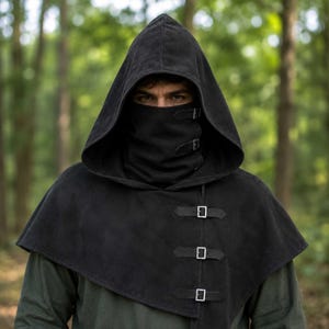 Peut inclure: Une capuche noire avec un cache-cou couvrant le visage et une cape arrivant aux épaules. Le vêtement est fixé par trois boucles métalliques argentées sur le devant du cache-cou et trois boucles assorties sur le devant de la cape.
