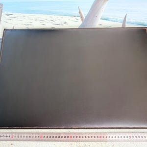 Peut inclure: Un sous-main rectangulaire marron foncé avec une surface texturée et des coutures visibles sur les bords. Une règle métallique placée le long du côté gauche et du bord inférieur indique que le tapis mesure environ 60 cm sur 40 cm.