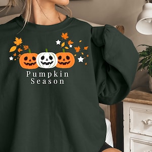 Könnte beinhalten: Ein dunkelgrüner Rundhals-Pullover mit einer Grafik von drei Kürbissen, zwei orangefarbenen und einem weißen, umgeben von fallenden orangefarbenen Blättern und weißen Sternen. Der Text "Pumpkin Season" ist in Weiß unter den Kürbissen gedruckt.