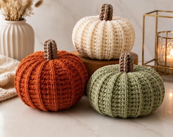 May include: Three crocheted pumpkins in orange, cream, and sage green. Each pumpkin features a ribbed texture and a brown crocheted stem. The pumpkins are arranged together, showcasing their rounded shapes and detailed yarn construction.