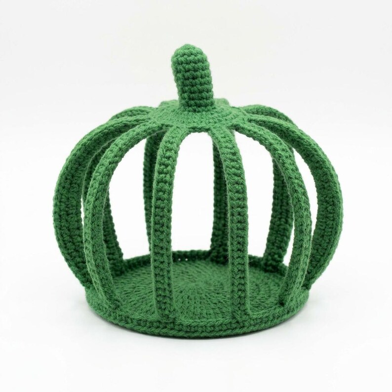 May include: A green crochet crown featuring a circular base and vertical arched ribs that meet at a central top point. The structure is crafted from yarn with a visible stitch pattern, creating a hollow, cage-like form with a solid circular bottom.