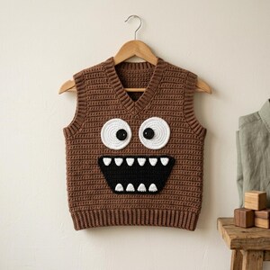 May include: A brown sleeveless V-neck sweater vest made of crochet yarn. The front features a large monster face design with two white circular eyes with black pupils and a wide black mouth containing several white triangular teeth.