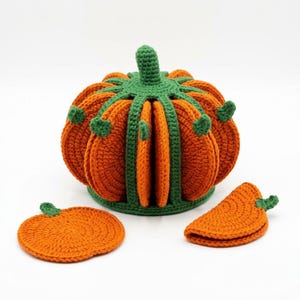 May include: A crochet pumpkin-shaped holder containing several orange circular coasters with green stems. The holder features a green ribbed structure that keeps the coasters upright. Two additional orange pumpkin-shaped coasters are placed in front.