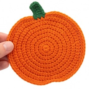 May include: A round, orange crocheted coaster shaped like a pumpkin, featuring a textured spiral stitch pattern and a small, dark green crocheted stem attached to the top edge. The item is crafted from yarn with a visible, consistent knit texture.