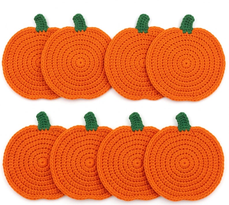May include: A set of eight circular orange crochet coasters, each featuring a small green stem at the top. The coasters are arranged in two rows of four, showcasing a consistent round, textured stitch pattern throughout the orange pumpkin-shaped bodies.