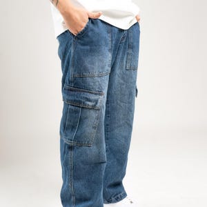 Könnte beinhalten: Blaue Jeans-Cargohose mit mehreren Taschen. Die Hose hat eine lockere Passform und wird mit weißen Turnschuhen kombiniert. Das Bild ist vor einem weißen Hintergrund aufgenommen, der die Details der Jeans hervorhebt.