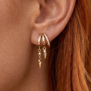 Könnte beinhalten: Drei goldene Creolen, die an einem Ohr getragen werden. Die beiden äußeren Ringe sind mit kleinen klaren Steinen besetzt, während der mittlere Ring einen hängenden Anhänger mit einem kleinen klaren Stein und einer goldenen Spitze aufweist.