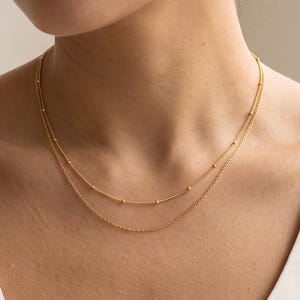 May include: A double-layered gold necklace featuring a delicate chain with small, evenly spaced gold beads on the upper strand and a simple, fine gold chain on the lower strand, resting against a plain white sleeveless top.
