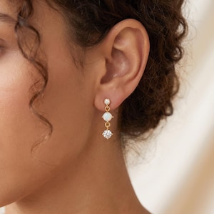May include: A gold-toned drop earring featuring three vertically aligned stones. The top stone is a small clear crystal, the middle is a round white opal, and the bottom is a larger clear crystal. The stones are set in gold metal links.