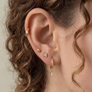 May include: A gold ear stack featuring a beaded cartilage hoop with a single clear stone, a small gold ball stud, a three-stone cluster stud, and a gold hoop earring with a dangling rectangular baguette-cut clear stone charm.