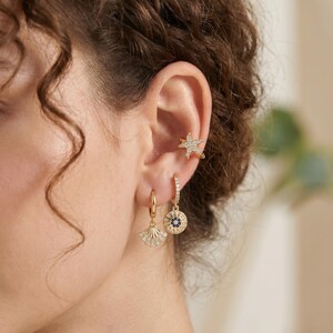 Può includere: Un ear cuff dorato a forma di stella marina incastonato con pietre chiare, abbinato a due orecchini a cerchio dorati. Un cerchio presenta un ciondolo a ventaglio con pietre chiare, l'altro un ciondolo circolare con una pietra blu centrale.