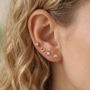May include: A set of four gold-toned stud earrings arranged along the earlobe. The designs include a four-pointed star with a central stone, a crescent moon with a small stone, a sunburst shape with a central stone, and a planet with a ring.