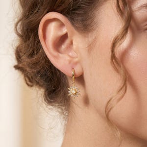 Peut inclure: Une boucle d'oreille créole dorée avec un pendentif en forme d'étoile. Le pendentif est orné de petits cristaux transparents et d'une pierre blanche ronde centrale. La boucle est fixée au lobe, révélant une finition dorée et un travail du métal.