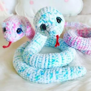 Peut inclure: Serpents crochetés à la main, dans des tons de bleu, blanc et rose. Les serpents ont de grands yeux noirs et des langues rouges. L'un des serpents est enroulé, l'autre est en position haute. La texture douce et moelleuse est visible.