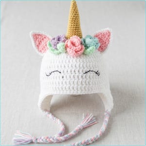 Puede incluir: Un gorro de unicornio blanco tejido a crochet con un cuerno dorado, orejas forradas en rosa y una corona floral de flores rosas, moradas y verdes. El gorro presenta una cara sonriente con ojos bordados en negro y lazos trenzados con borlas pastel.
