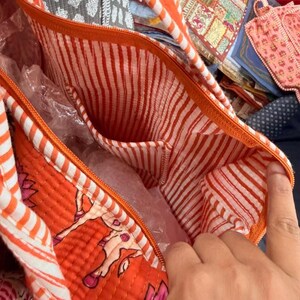 May include: A quilted tote bag featuring an orange exterior with a floral pattern and a white and orange striped interior lining. The bag includes an orange zip closure and an integrated internal pocket with the same striped fabric design.