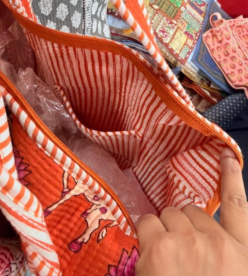 May include: A quilted tote bag featuring an orange exterior with a floral pattern and a white and orange striped interior lining. The bag includes an orange zip closure and an integrated internal pocket with the same striped fabric design.