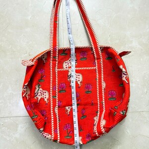 May include: A red quilted tote bag featuring a repeating pattern of white cows and pink lotus flowers. The bag has striped handles and trim. A white measuring tape is placed vertically over the centre, showing a length of approximately 51 cm.