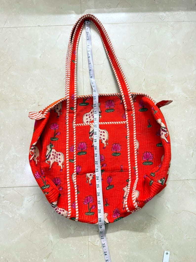 May include: A red quilted tote bag featuring a repeating pattern of white cows and pink lotus flowers. The bag has striped handles and trim. A white measuring tape is placed vertically over the centre, showing a length of approximately 51 cm.