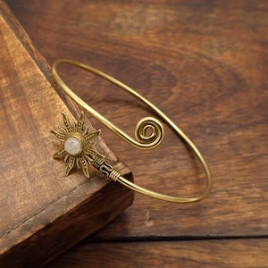 May include: A gold-toned metal cuff bracelet featuring a sun-shaped charm at one end with a central white stone and a spiral design at the opposite end. The bracelet has a smooth, polished finish and an open, adjustable band structure.