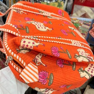 May include: An orange quilted bag featuring a repeating pattern of cream-coloured cows with pink spots and pink lotus flowers on green stems. The bag includes white and orange striped fabric trim along the edges and a silver metal zip closure.