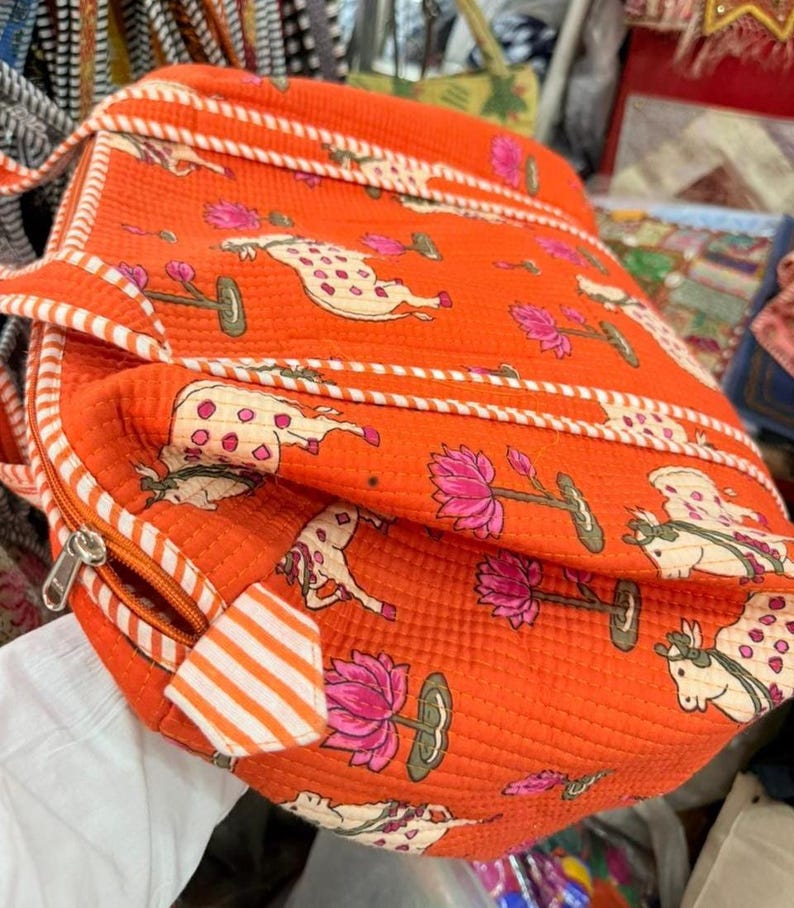 May include: An orange quilted bag featuring a repeating pattern of cream-coloured cows with pink spots and pink lotus flowers on green stems. The bag includes white and orange striped fabric trim along the edges and a silver metal zip closure.